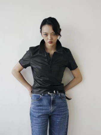 Adult woman in a black leather polo shirt and jeans stands in a studio setting, showcasing fashion apparel against a plain background.