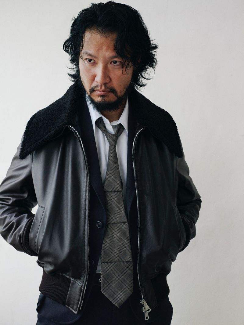 Portrait of a stylish Asian man in a dark leather bomber jacket, suit, and tie against a neutral background in a studio setting.
