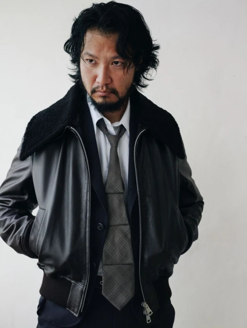 Portrait of a stylish Asian man in a dark leather bomber jacket, suit, and tie against a neutral background in a studio setting.