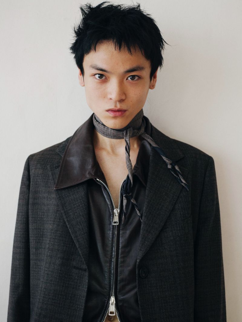 Headshot of an androgynous young adult with black hair wearing a jacket, silky shirt, and patterned scarf, against a white background.