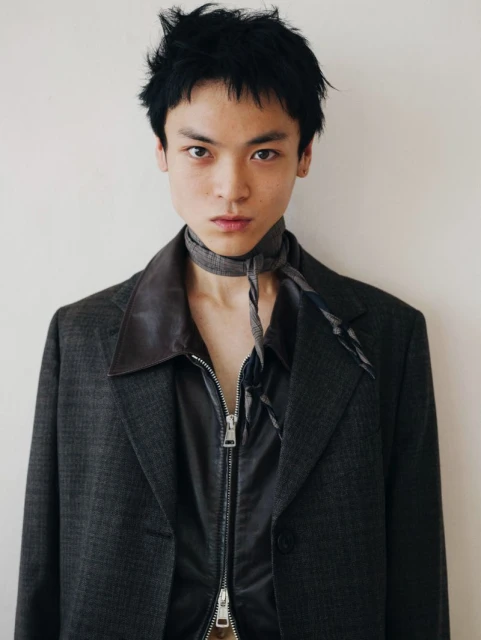 Headshot of an androgynous young adult with black hair wearing a jacket, silky shirt, and patterned scarf, against a white background.