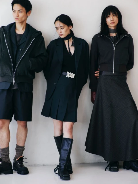 Three Asian models in an indoor studio setting showcasing dark-colored fashion. A hooded jacket, skirt, and long dress are featured.