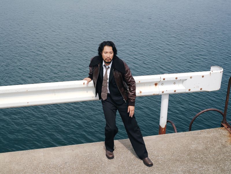 Adult male in suit jacket and tie poses on a pier near dark ocean water and cloudy skies with a serious expression on his face.