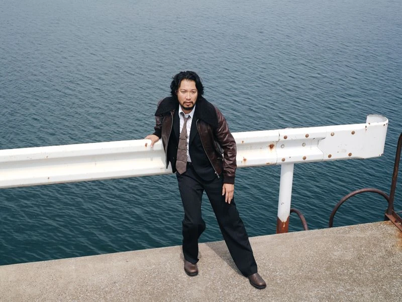 Adult male in suit jacket and tie poses on a pier near dark ocean water and cloudy skies with a serious expression on his face.