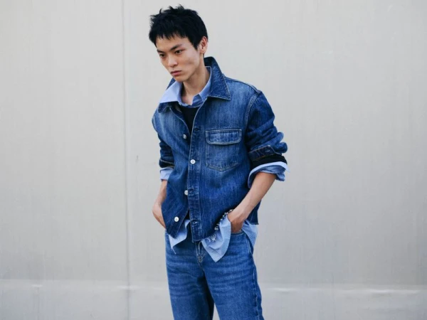 Young man in a denim jacket and blue jeans standing against a plain pale wall