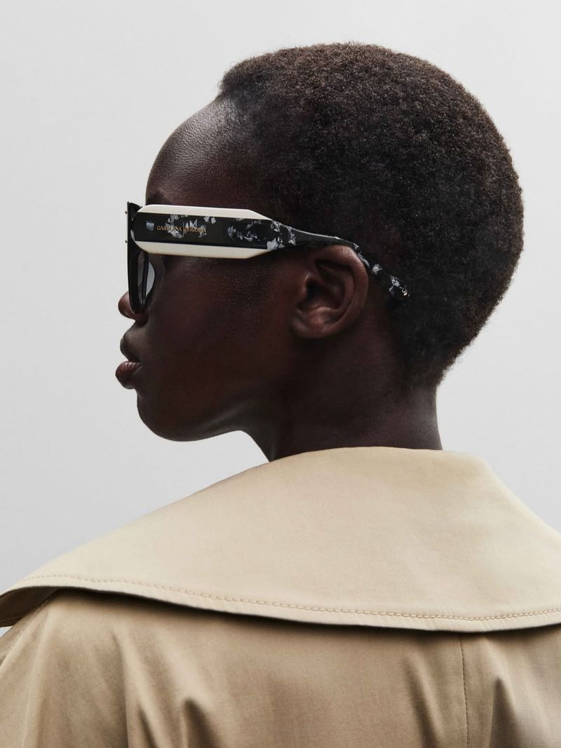 Side profile of a Black woman wearing black and white sunglasses and a beige trench coat against a neutral background.