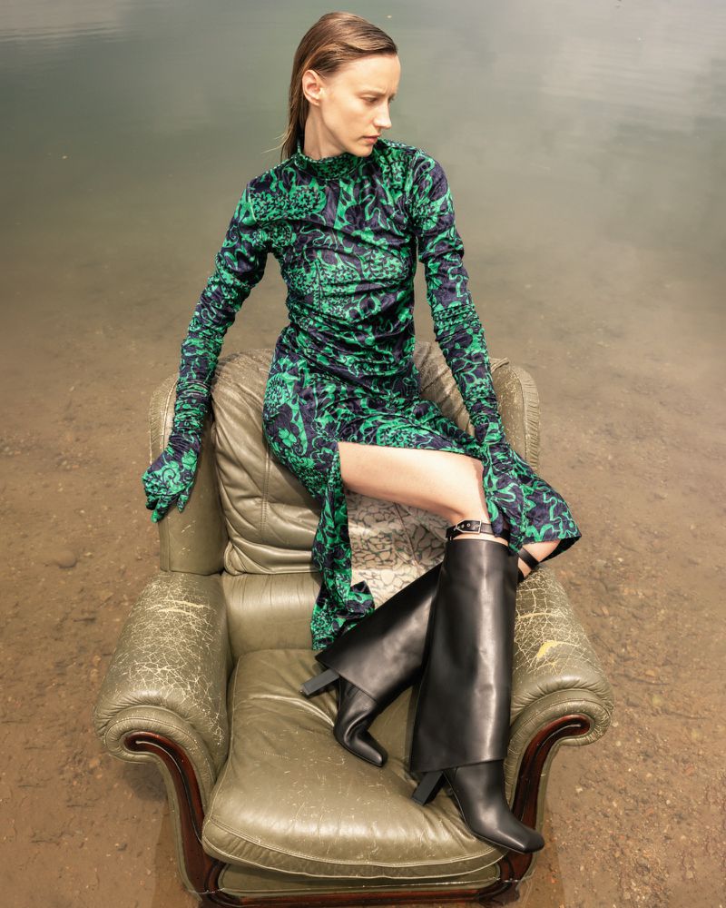 Woman in a green printed dress and black boots sits in a vintage armchair in a shallow lake, creating a surreal fashion editorial.