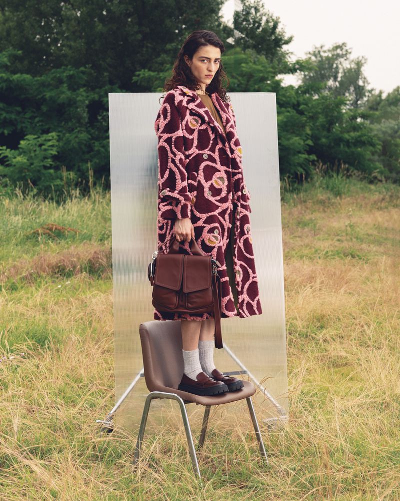 A woman in a patterned coat poses next to a clear panel in a field, sitting on a chair and holding a satchel-style purse.