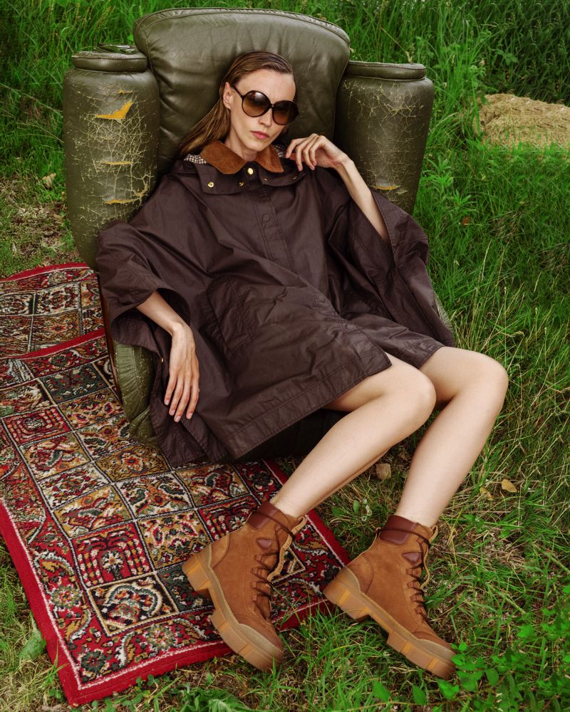 Woman in brown poncho, sunglasses, and boots reclines in armchair on an oriental rug in a green grassy outdoor setting on a sunny day.