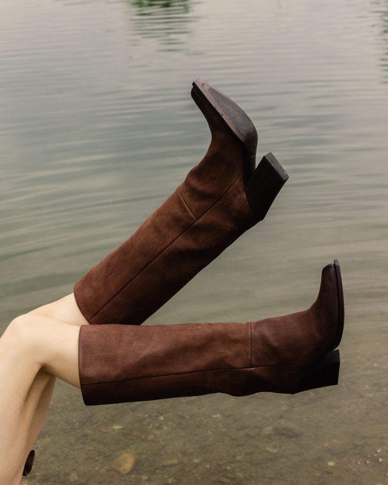 Brown leather knee-high boots are featured against an out-of-focus water background. The boots are the primary focus of the frame.