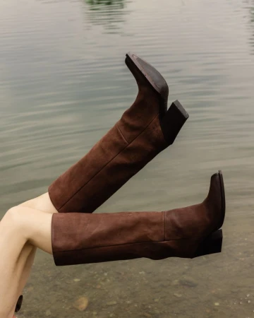 Brown leather knee-high boots are featured against an out-of-focus water background. The boots are the primary focus of the frame.