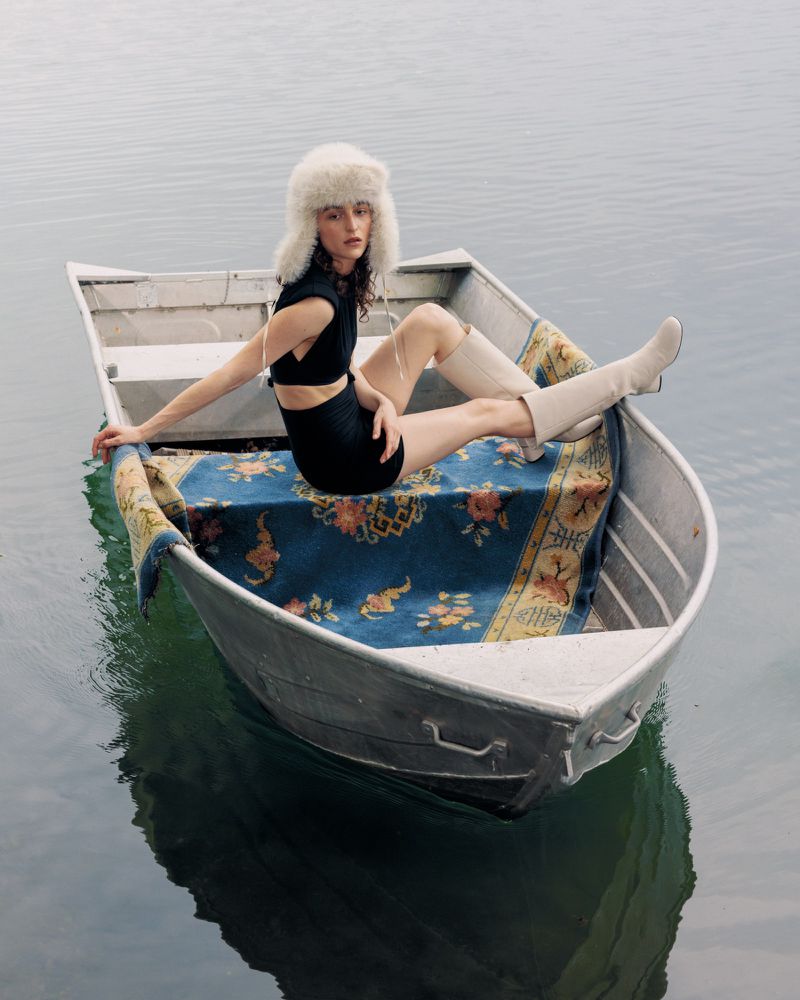 Woman in a black dress and furry hat sits in a metal rowboat on calm water, posing on a blue oriental rug on the boat's floor.