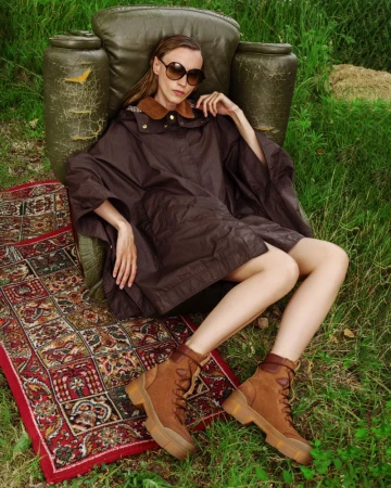 Model wearing a brown cape jacket and tan lace-up boots sitting in a weathered armchair on a patterned rug outdoors.