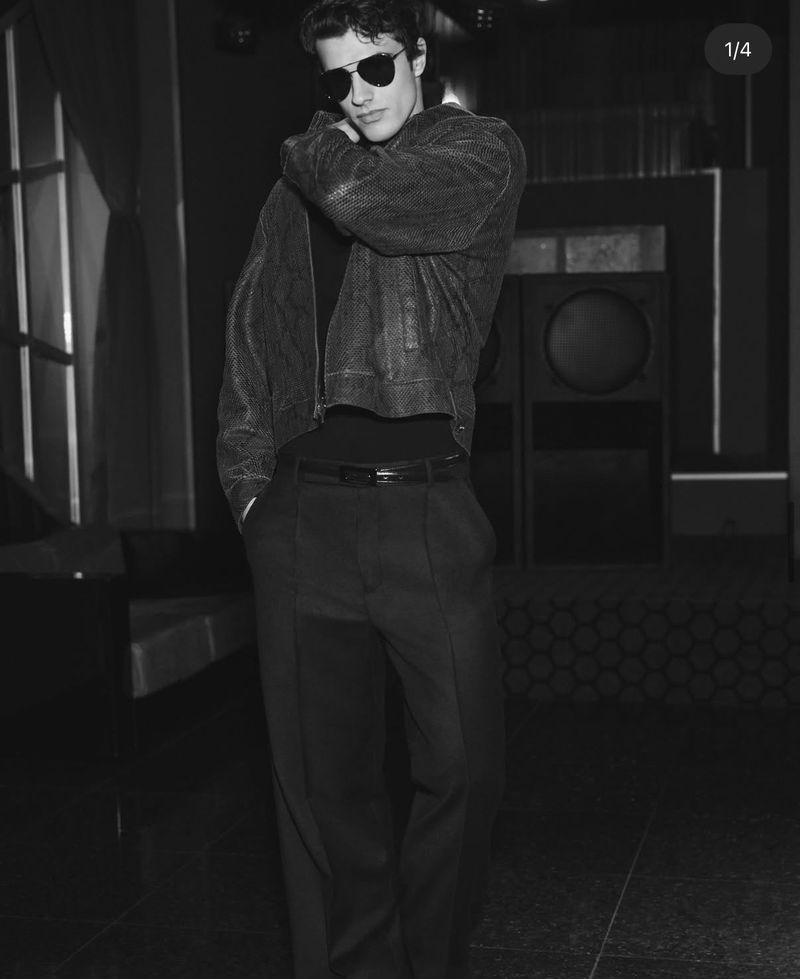 Monochrome studio shot of a young adult male model with sunglasses wearing a jacket and dress pants looking off camera.