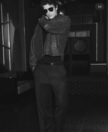 Monochrome studio shot of a young adult male model with sunglasses wearing a jacket and dress pants looking off camera.