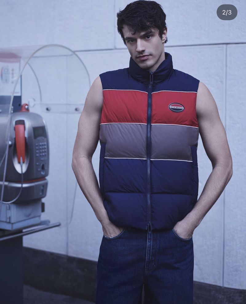 Man in blue jeans and a multi-colored puffer vest posing indoors next to a retro telephone booth with a light blue background.