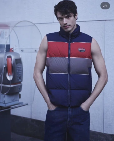 Man in blue jeans and a multi-colored puffer vest posing indoors next to a retro telephone booth with a light blue background.