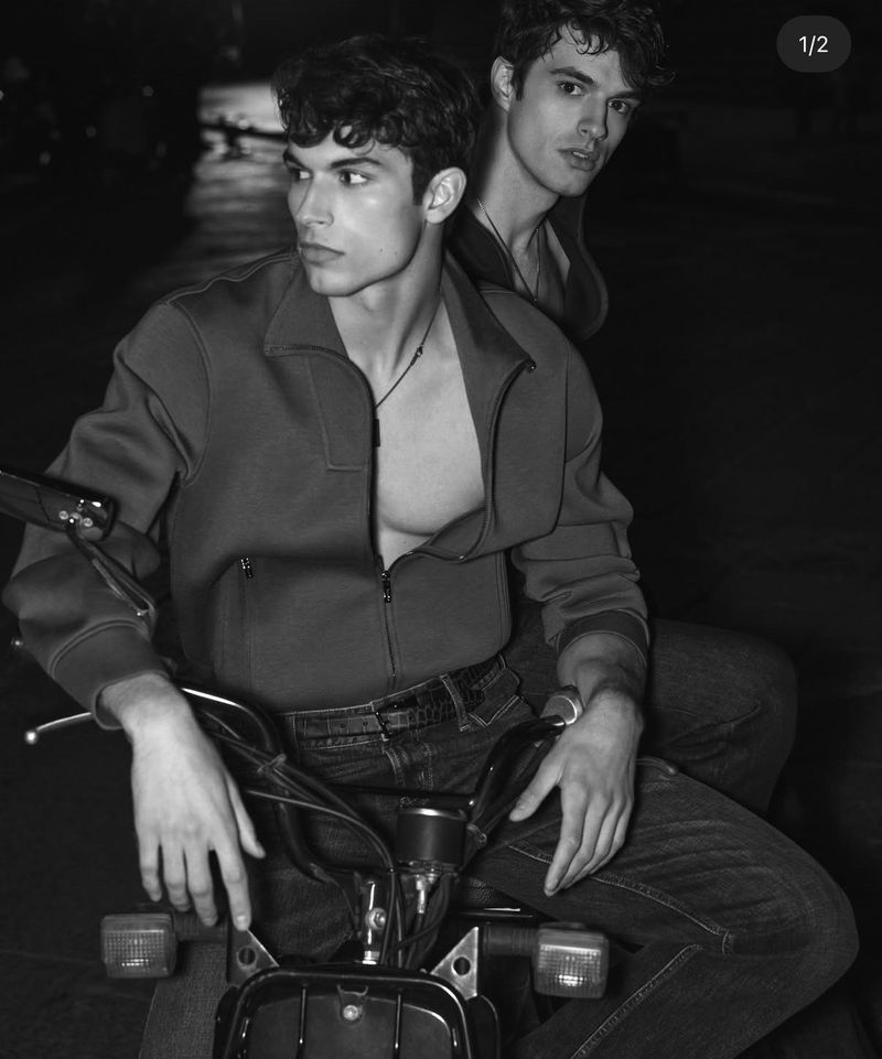 Two adult men sit on a black motorcycle, wearing jackets and jeans in a black and white outdoor shot at nighttime.