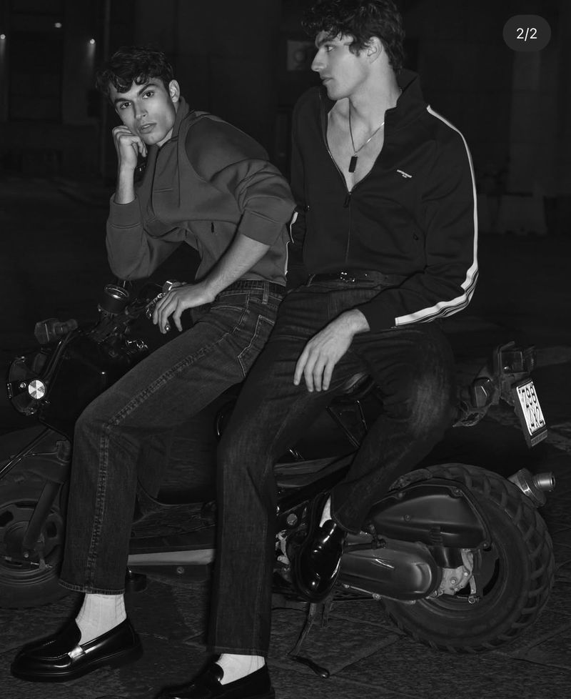 Two adult men wearing jackets and jeans sit on a scooter in a black and white nighttime fashion editorial with outdoor setting.