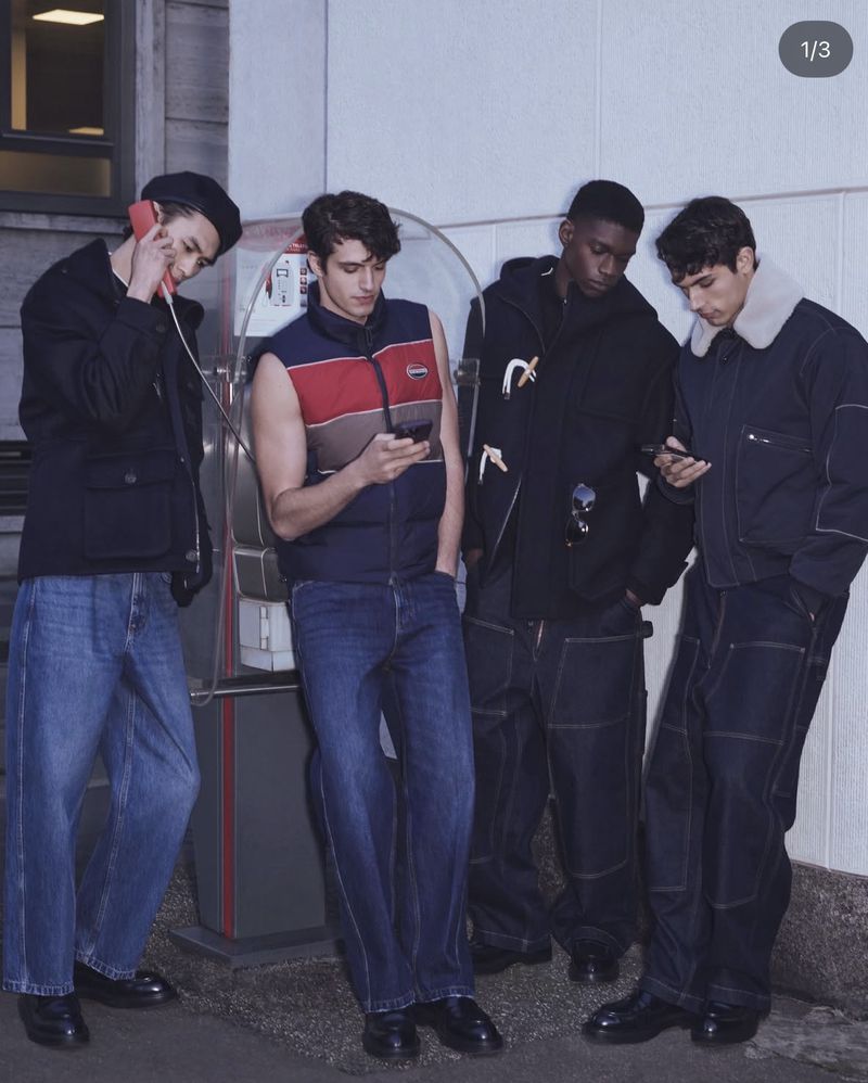 Four young adult men in wide leg jeans stand near a concrete column in an urban setting, one using a public telephone and the others using phones.