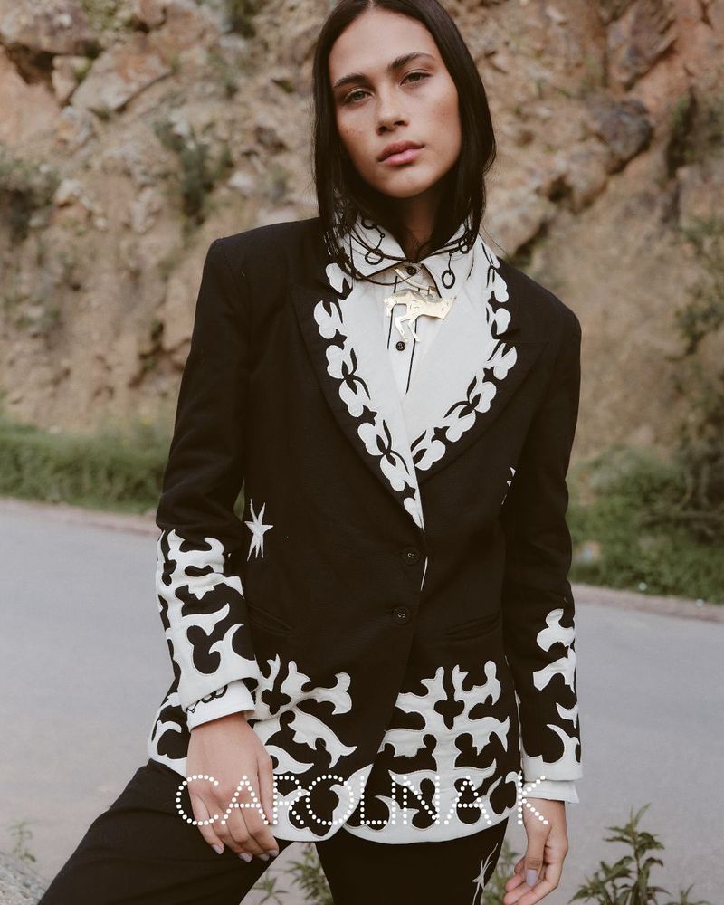 An adult female model poses in a black and white patterned suit and shirt, set against an outdoor backdrop of neutral tones.