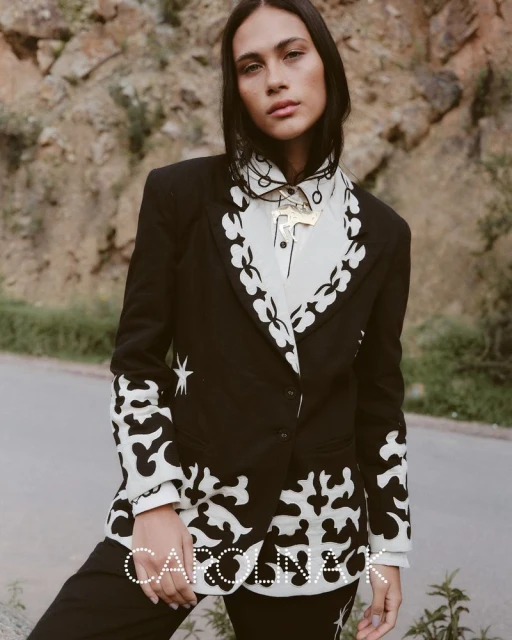 An adult female model poses in a black and white patterned suit and shirt, set against an outdoor backdrop of neutral tones.