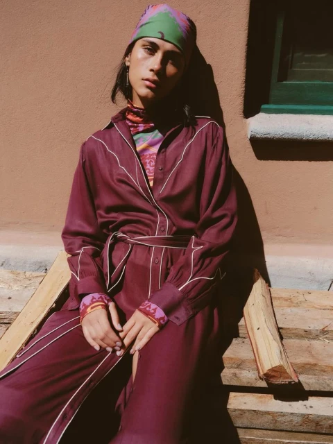 A person sits on a stack of wood wearing a long belted dress with a patterned headscarf, posing against a terracotta wall.