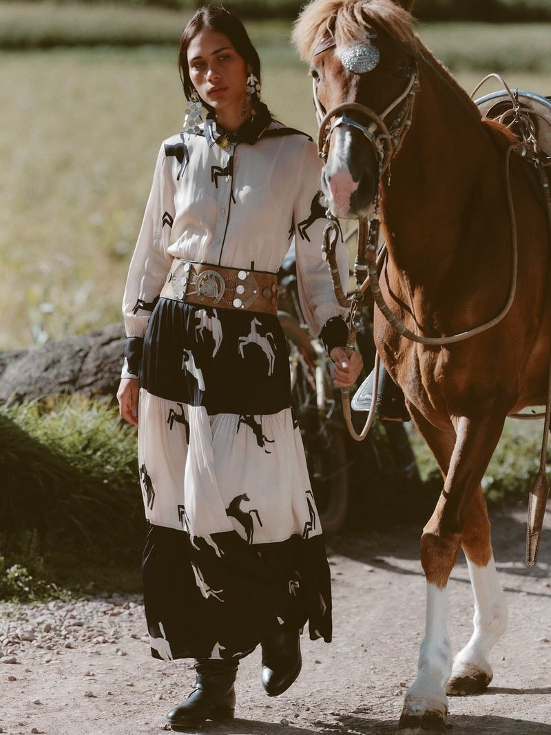 Fashionable woman in a horse-print dress stands next to a chestnut horse in a rural setting, showcasing a blend of elegance and country style.