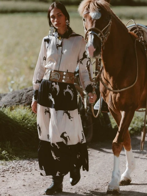 Fashionable woman in a horse-print dress stands next to a chestnut horse in a rural setting, showcasing a blend of elegance and country style.
