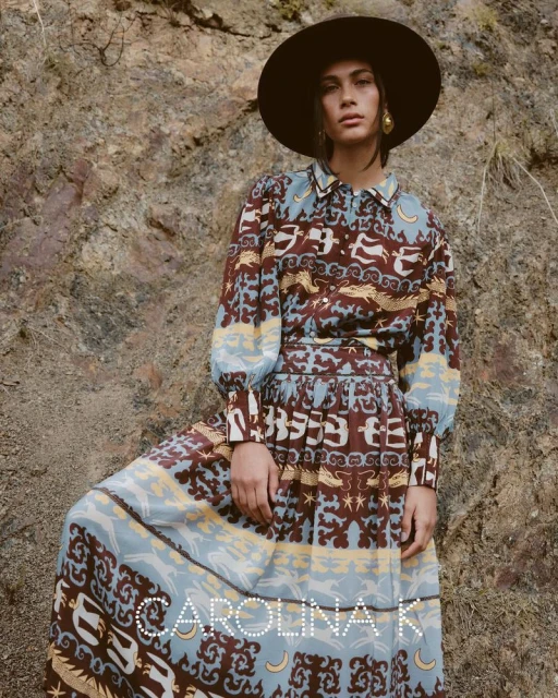 A stylish woman poses in a patterned dress and wide-brimmed hat against a rocky backdrop, showcasing an elegant, earthy fashion aesthetic.