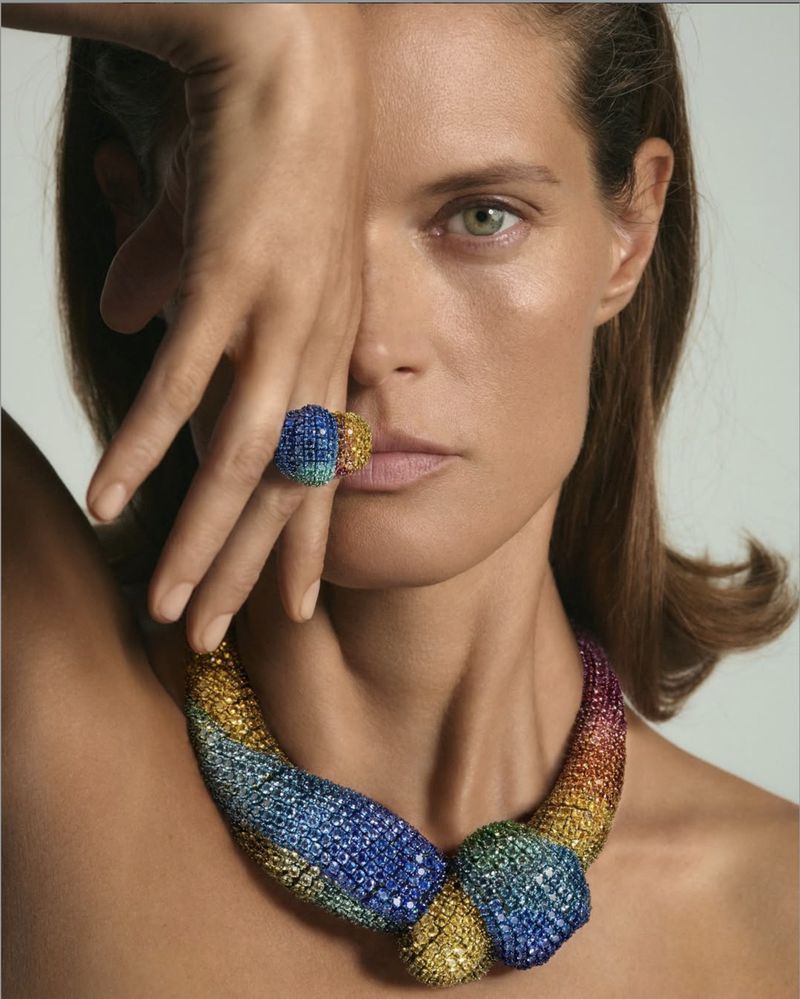 Close-up shot of a model wearing an elaborate multicolored ring and necklace made of small stones, against a pale backdrop.