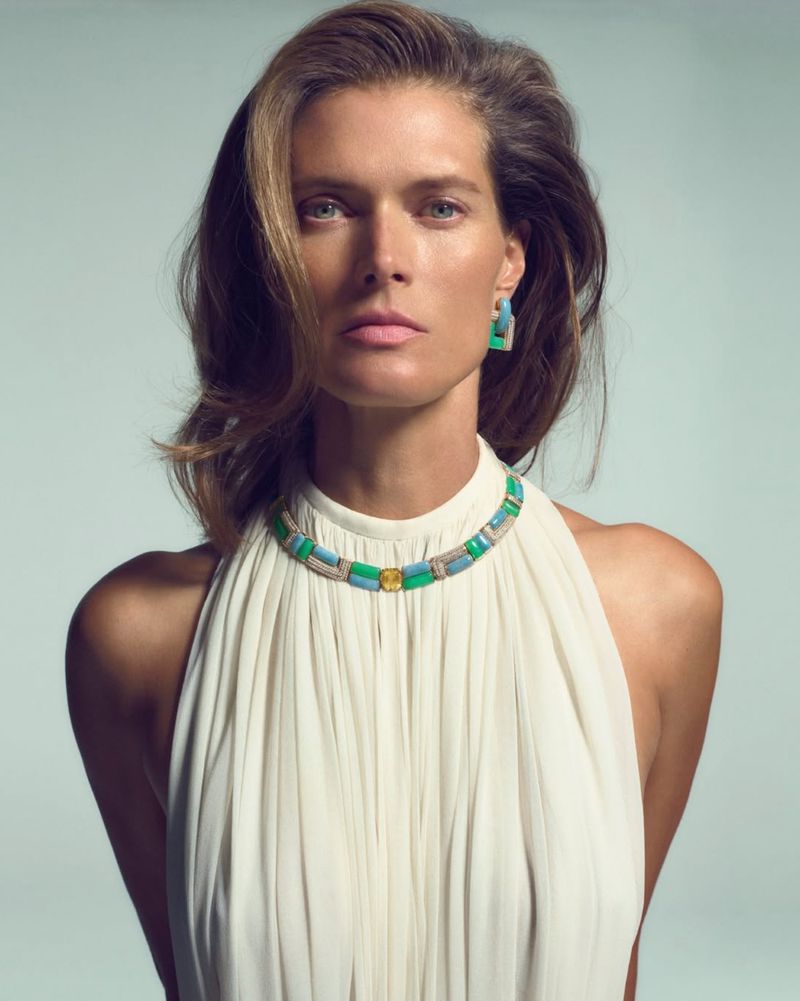 Portrait of an adult woman wearing a sleeveless white dress and colorful gemstone jewelry, including a necklace and earrings, in studio lighting.