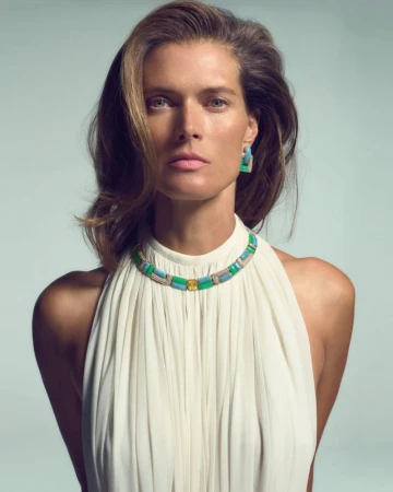 Malgosia Bela wearing a white pleated halter dress and a colorful Piaget gemstone necklace against a neutral studio backdrop.