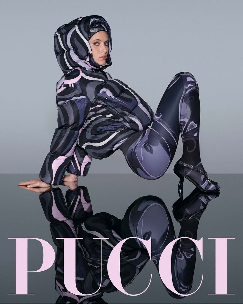 Woman poses in patterned puffer jacket and leggings with matching high heels on reflective surface, promoting fashion brand Pucci.