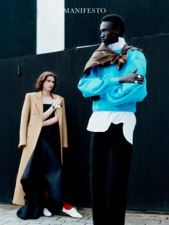 Two fashion models posing outdoors against a dark ribbed wall, one in a bright blue sweater and the other in a camel tailored coat.