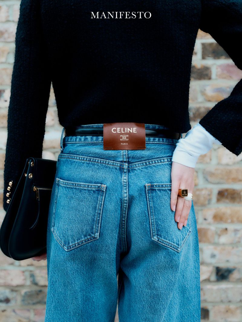 Close-up from behind shows blue jeans, black top, and leather Celine label detail against a brick wall, capturing fashion aesthetic.