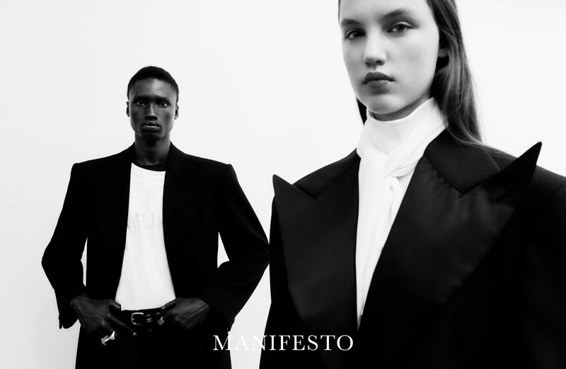 Black-and-white fashion shot featuring two models, an African-American man and a Caucasian woman, wearing black blazers in a studio setting.