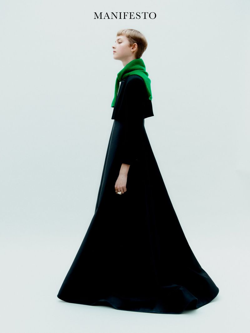 A slender woman with short hair poses sideways in a black, full-length dress with a bright green scarf against a bright white background.