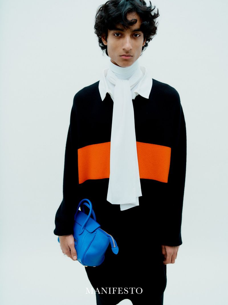 Editorial fashion shot of a young adult male in a black and orange sweater holding a blue handbag against a white backdrop in studio.