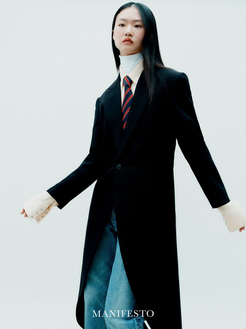 Asian woman in a long black coat, blue jeans, and tie against a white background in a fashion editorial style full studio shot.