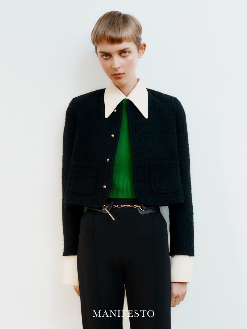 Woman in a black cropped jacket with gold buttons, a green sweater, and black pants against a white background in a studio setting.