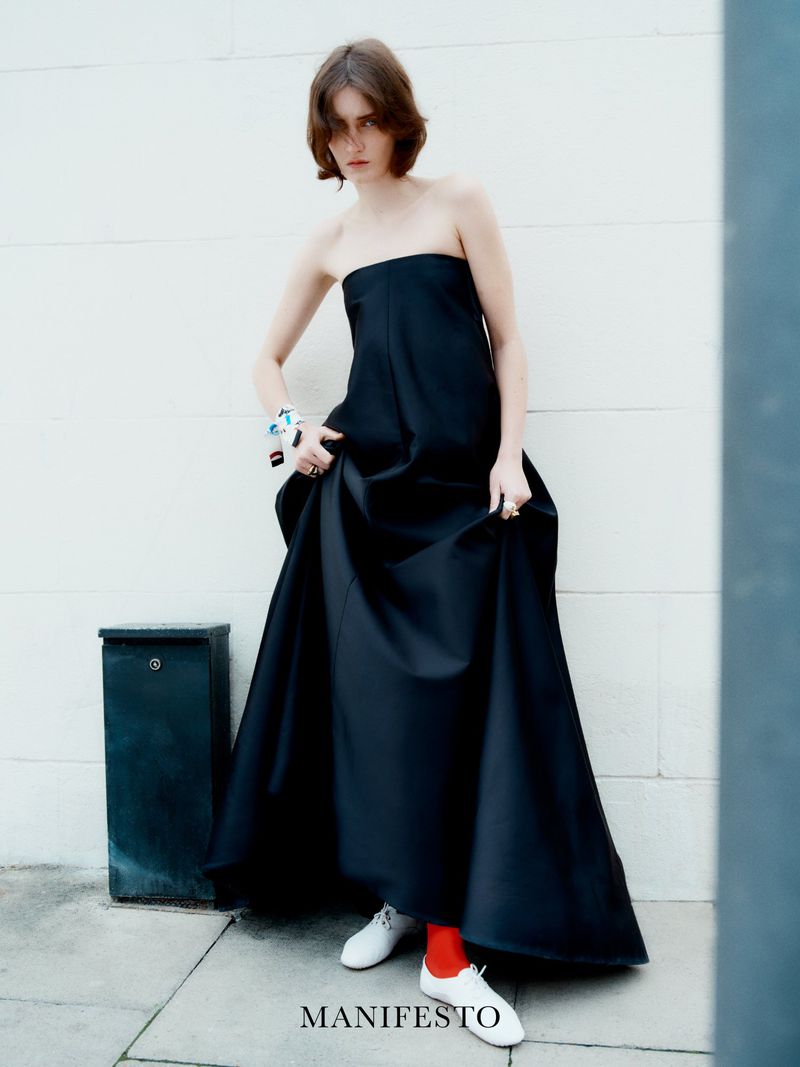 A female fashion model in a strapless black dress poses against a white wall, holding the dress hem, with red socks and white shoes.