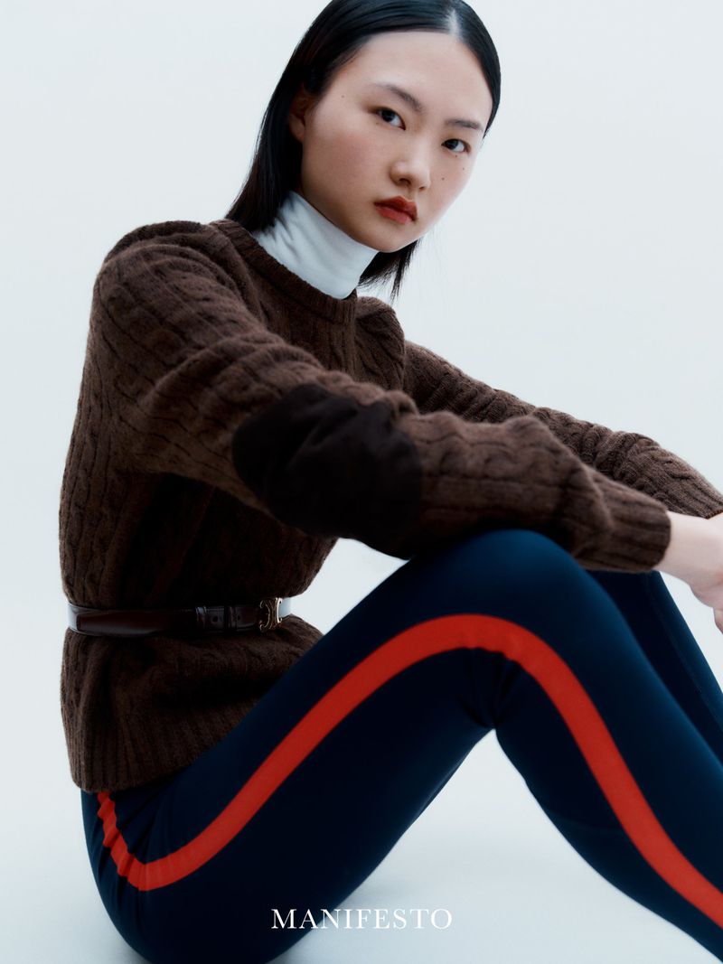 Asian woman in studio wearing a brown sweater and dark blue pants with a red stripe, seated in a bright, soft studio setting.