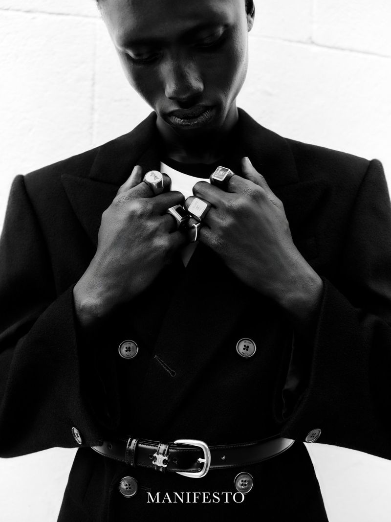 Black and white portrait of a Black man wearing a belted black double-breasted coat with silver rings holding his lapels.