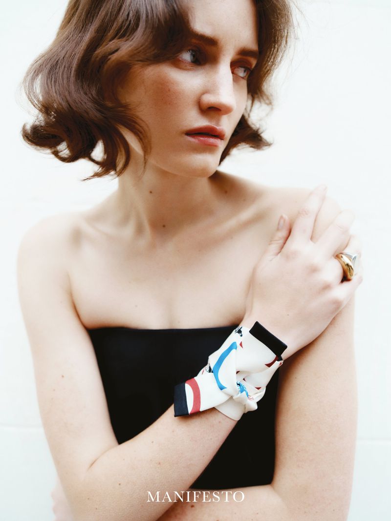 A woman with shoulder-length wavy brown hair wears a black strapless top and a patterned scarf tied around her wrist, posing in studio.