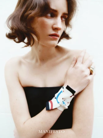 A woman with shoulder-length wavy brown hair wears a black strapless top and a patterned scarf tied around her wrist, posing in studio.