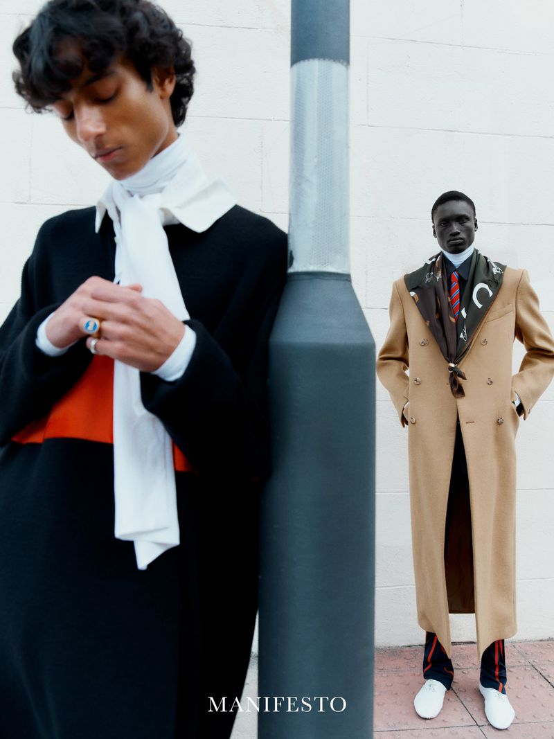 Two dark-skinned men pose outside in fashionable overcoats, one black and one tan, accessorized with white shoes and scarves.