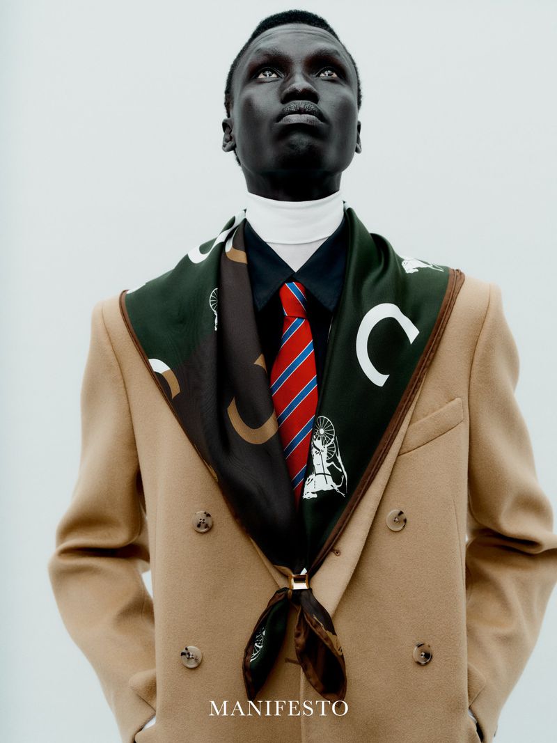 Man wearing camel coat, patterned green scarf, red striped tie and white turtleneck against plain background