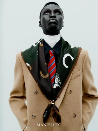 Man wearing camel coat, patterned green scarf, red striped tie and white turtleneck against plain background