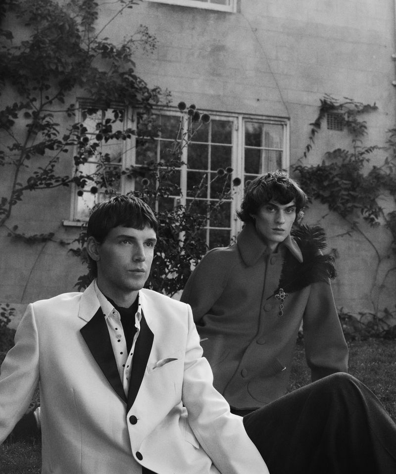 Two men pose in formal wear outside a building with climbing vines. The monochrome image features a white blazer and textured coat.